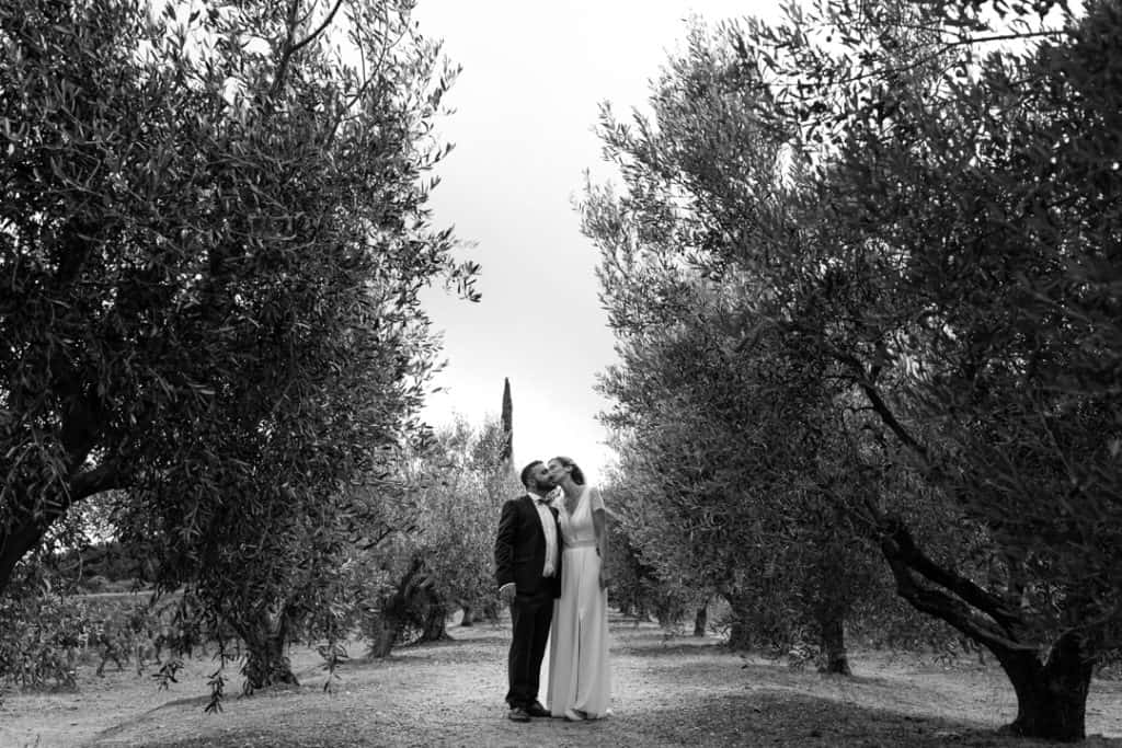 Un couple de mariés s'embrasse au milieu des oliviers. Photo en noir et blanc. 