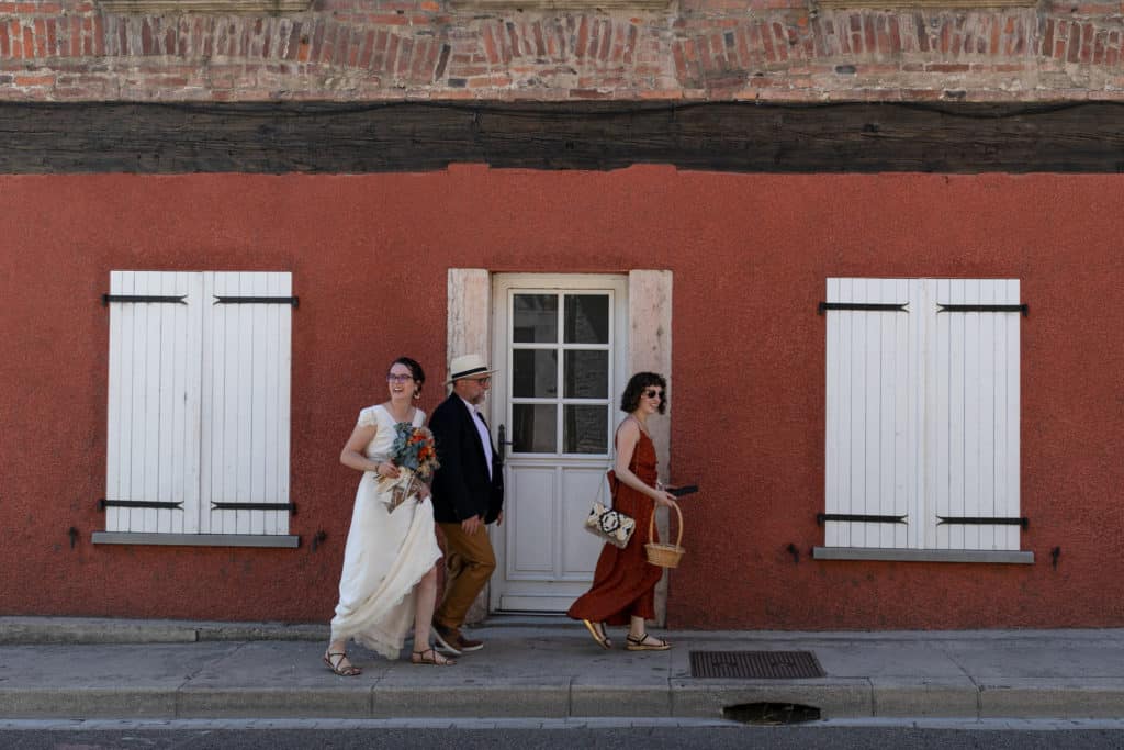 La mariée marche devant un mur rouge. 
Mariage Amandine et Antoine au domaine de Marze à Saint Georges de Reneins, Rhône.