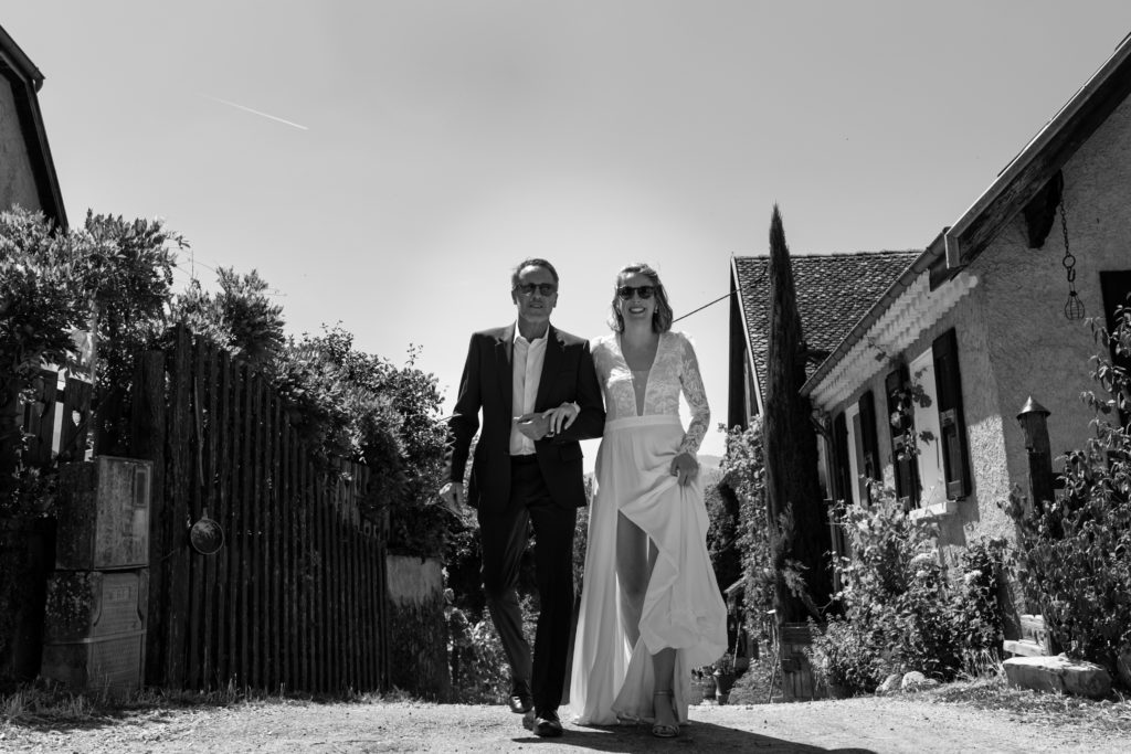 La mariée au bras de son père, marche en direction de la mairie. Mariage à Prébois, près de Grenoble, Région Rhône-Alpes. Photographie prise par Sabine Greppo, photographe de mariage à Lyon et en France.